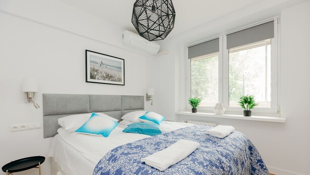 A neatly made bed with a blue patterned bedspread, a grey headboard, and a window with a view of greenery. - ShortStayPoland Jana Pawła - B27 (Warsaw)