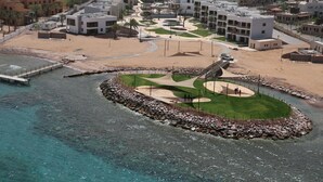 An aerial view of a coastal resort with modern buildings, a sandy beach, and a rocky water feature. - Public Security Hotel & Chalets (Aqaba)
