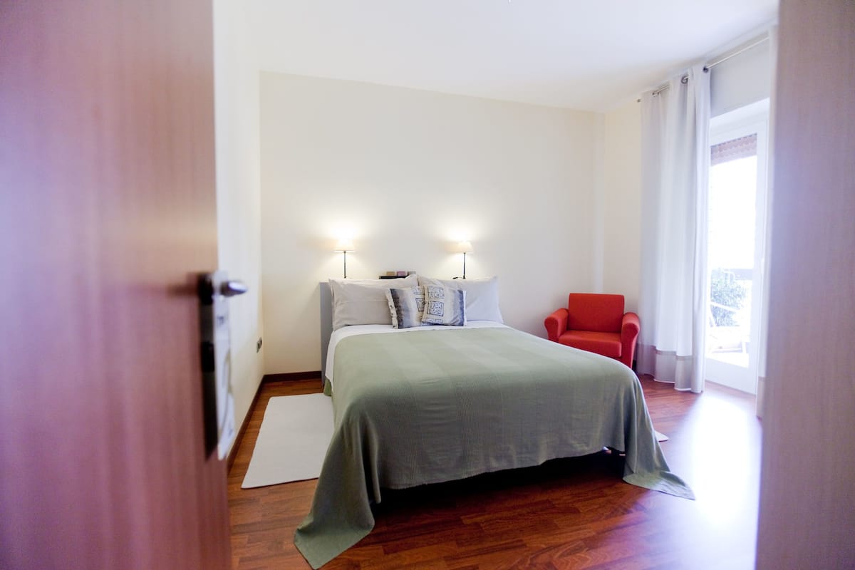 A bedroom with a bed, two bedside lamps, a red chair, and a large window with curtains.