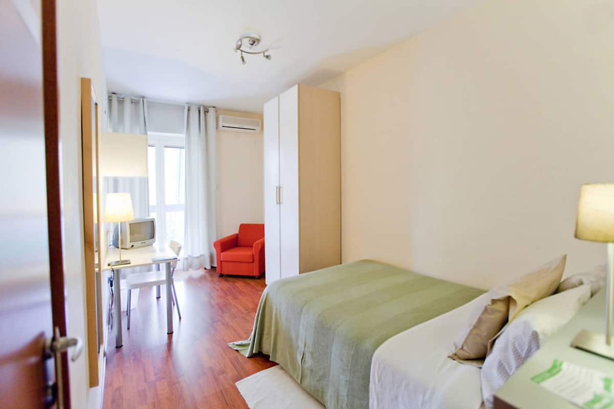 A hotel room with a bed, a desk with a computer, a chair, and a window with curtains.