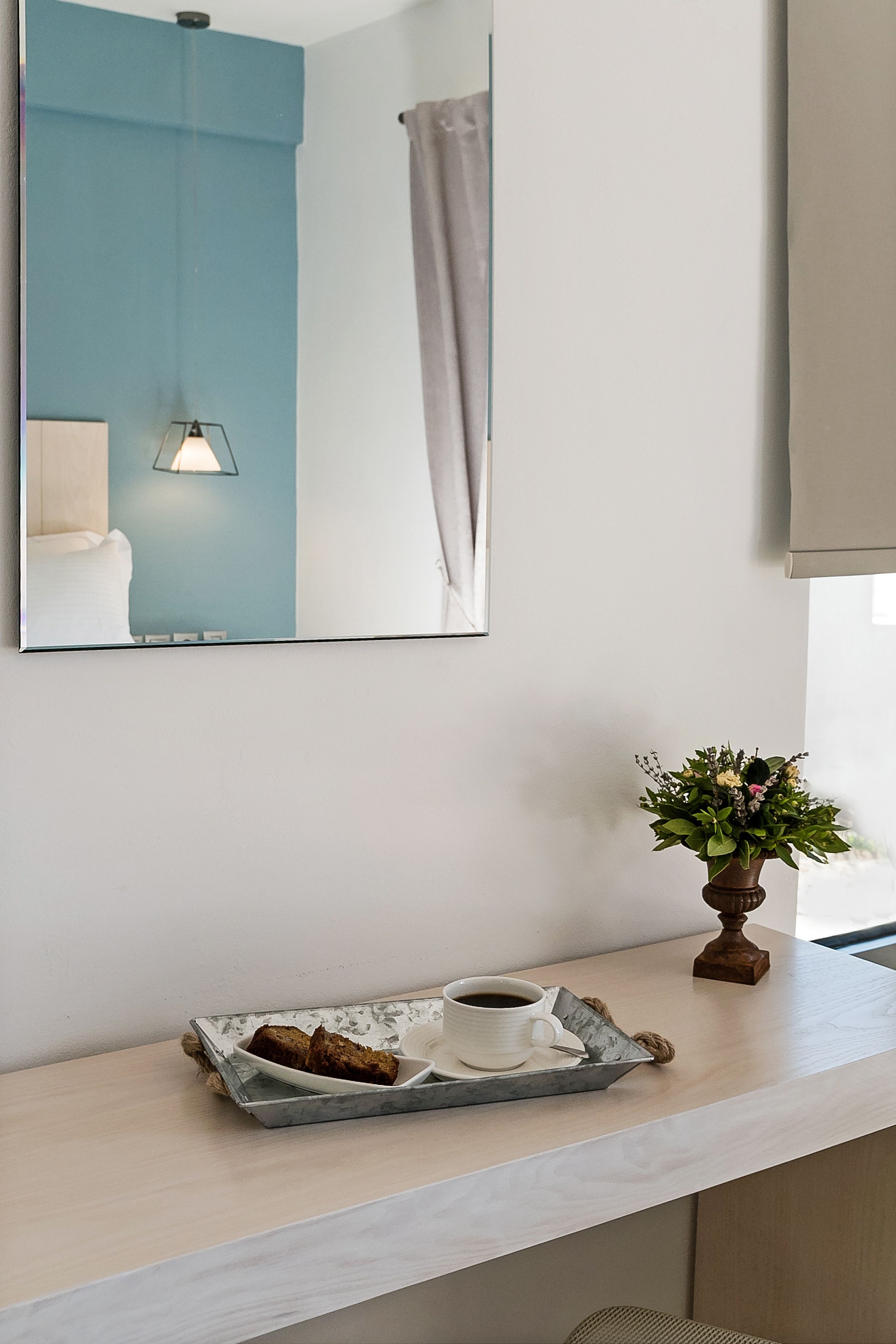 Habitación de hotel con una consola de mesa de color claro, una bandeja con una taza de café y una porción de pastel, una planta en maceta y un espejo grande que refleja una pared azul y una ventana con cortinas.