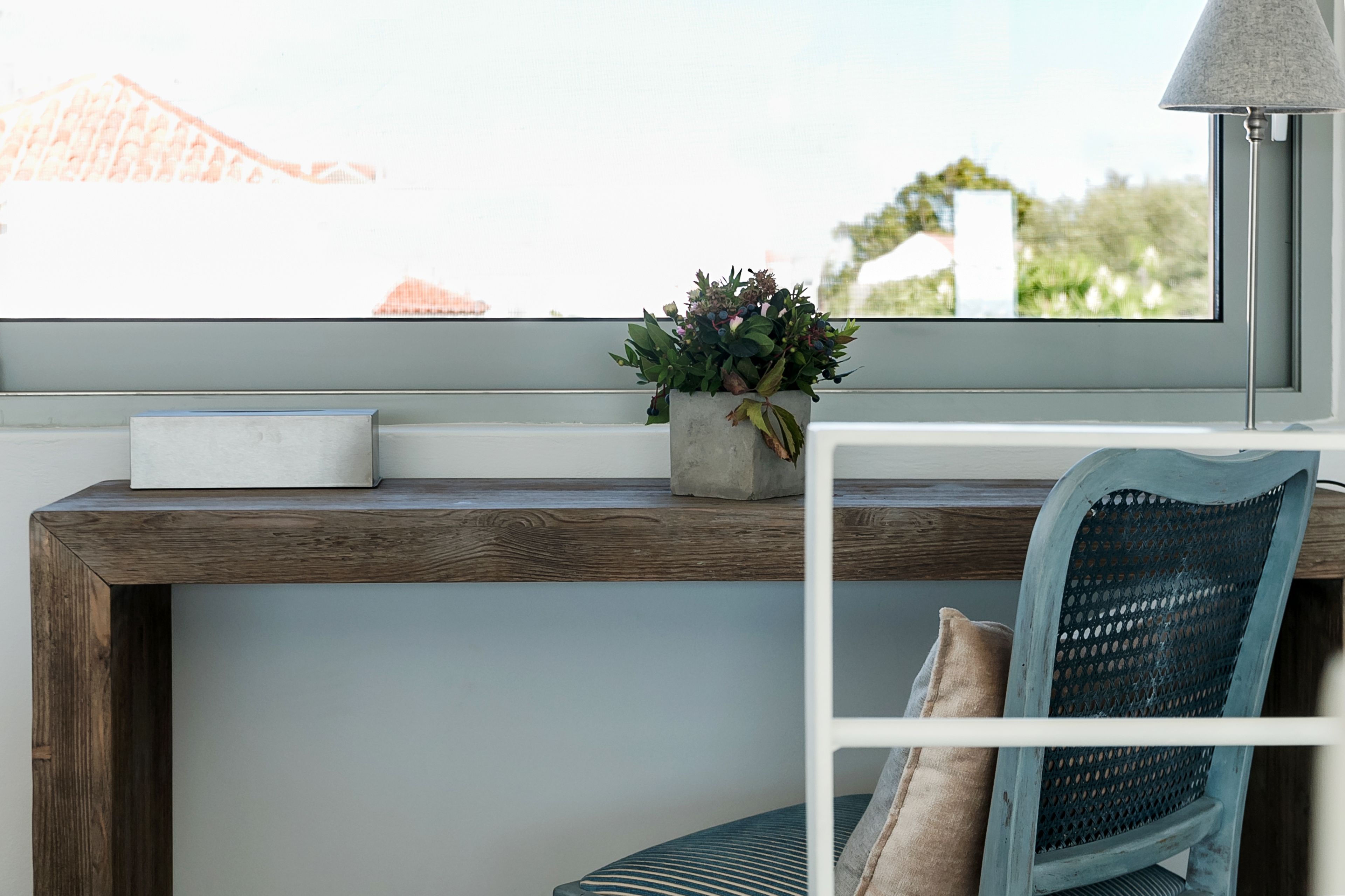A window with a view of buildings, a concrete planter with plants, a white lamp, and a chair with a woven backrest.