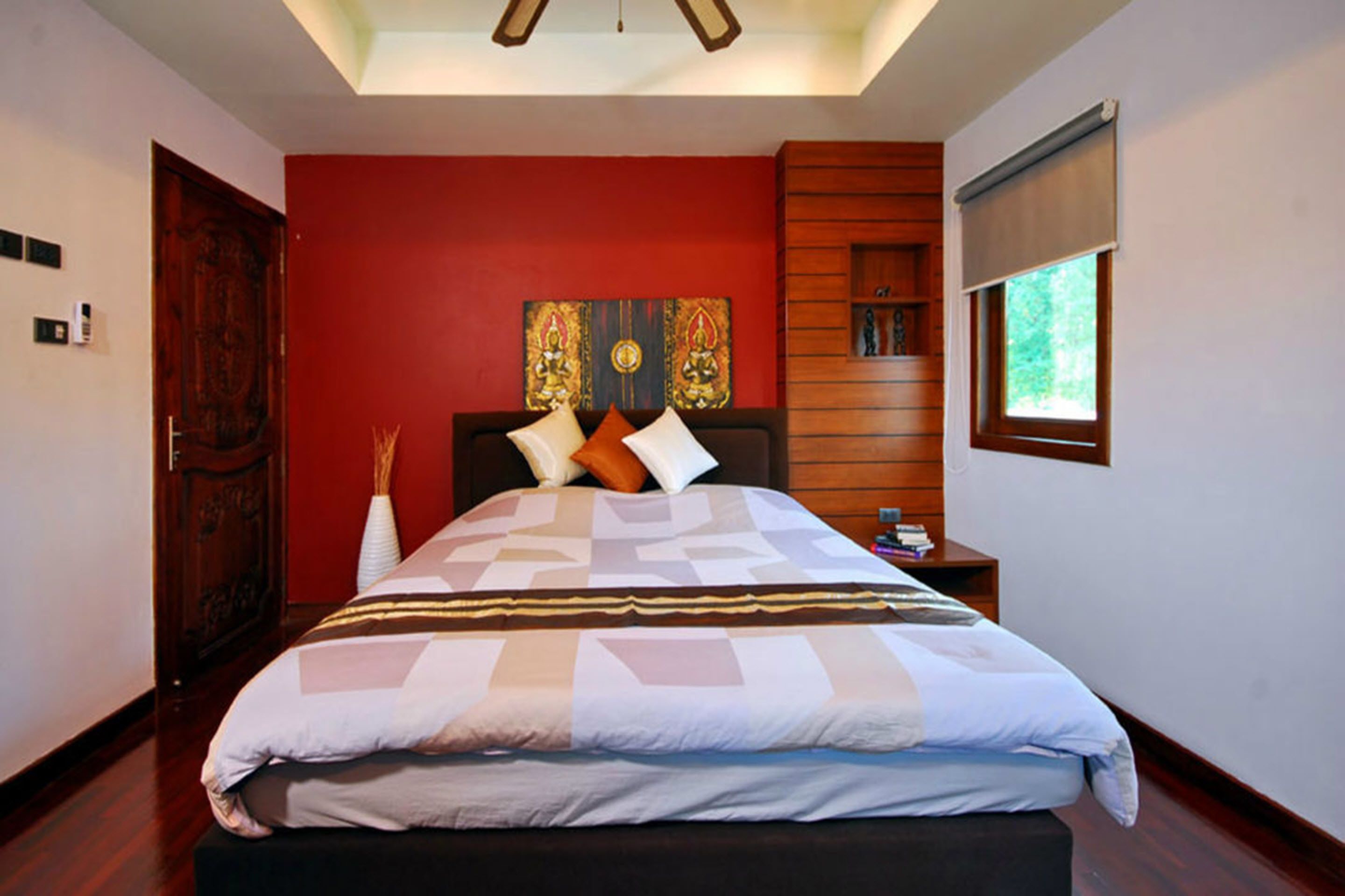 A bedroom with a bed, a wooden headboard, a red accent wall, a window with blinds, and a ceiling fan.