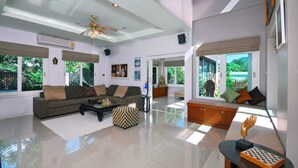 A spacious living room with a large sofa, a coffee table, and a checkered ottoman. There is a glass table with a chessboard and a golden hand sculpture. - 4 Bedroomed Villa In Chaweng P1 SDV193 - By Samui Dream Villas (Koh Samui)
