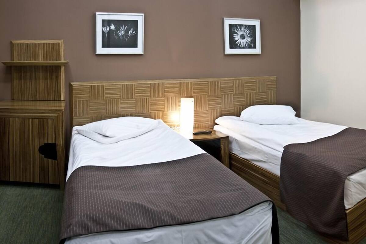 A hotel room with two single beds, a wooden headboard, a nightstand with a lamp, and two framed pictures on the wall.