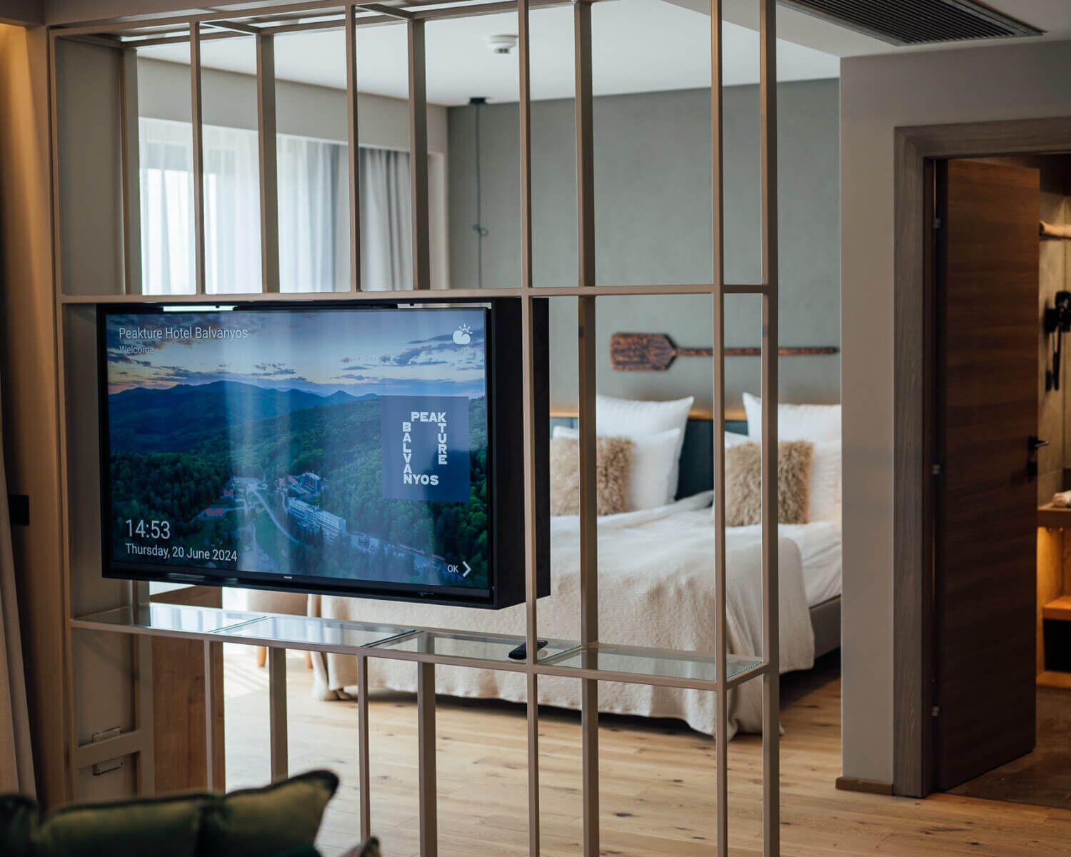 A hotel room with a bed, a TV mounted on a glass partition, and a wooden floor.