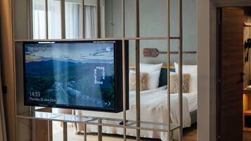A hotel room with a bed, a TV mounted on a glass partition, and a wooden floor.