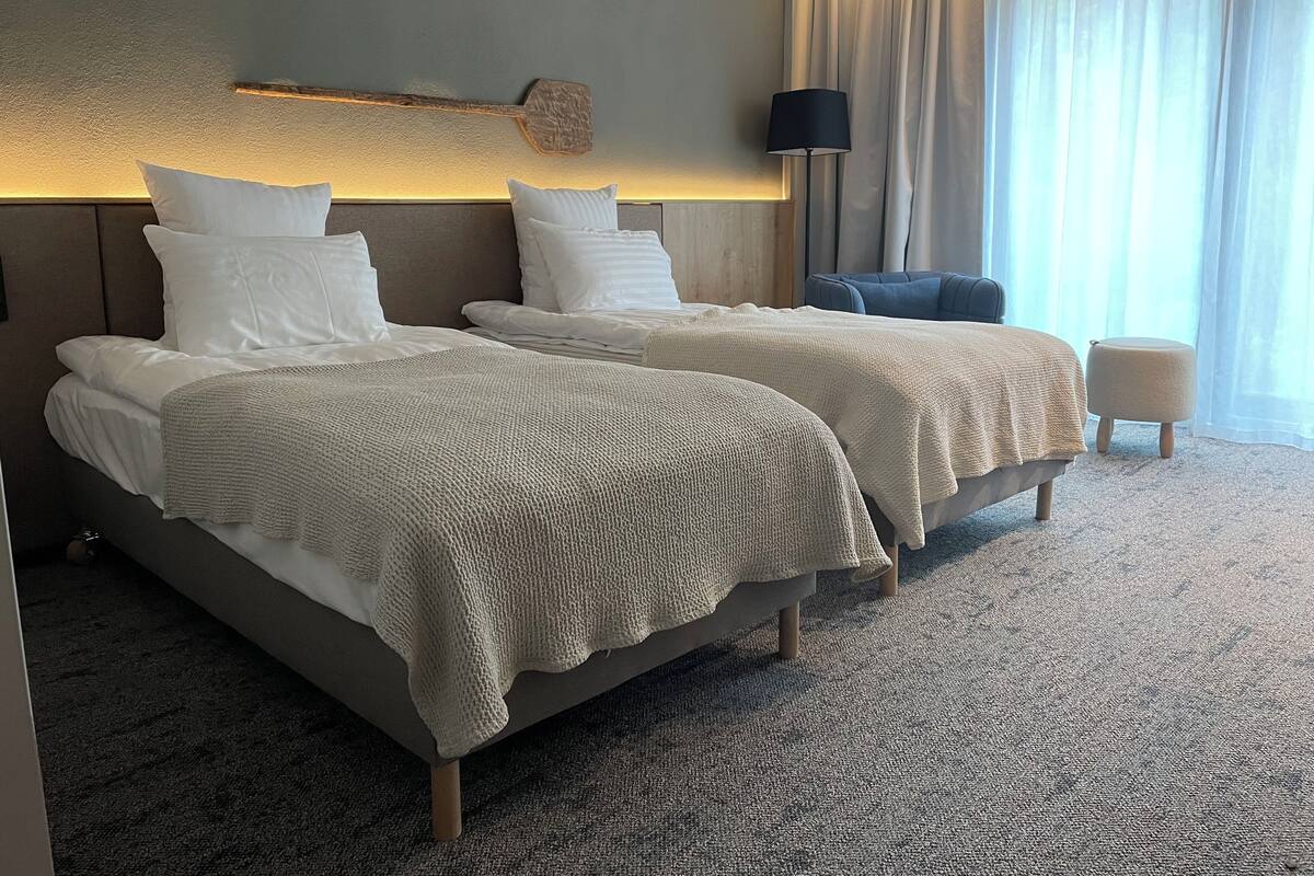 A hotel room with two beds, a nightstand, a lamp, and a view of the outdoors.