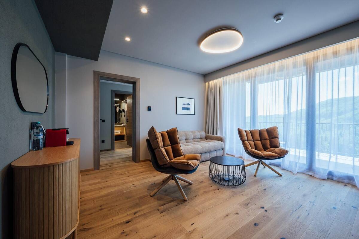 A modern living room with a wooden floor, two brown armchairs, a round coffee table, and a sofa. There is a large window with sheer curtains, a ceiling light, and a framed picture on the wall.