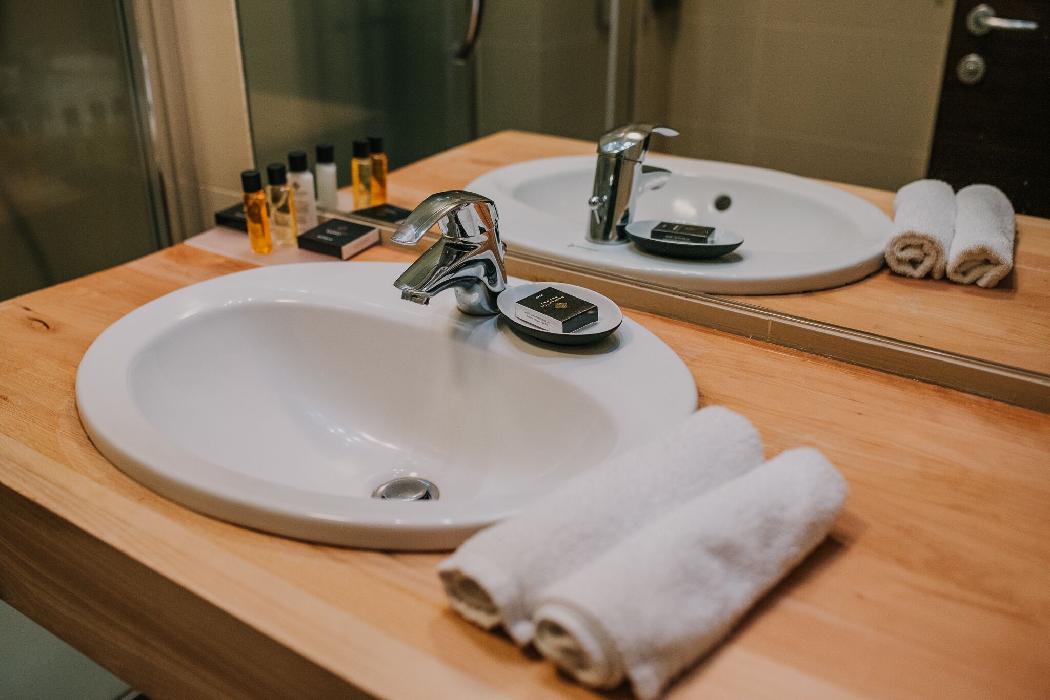 Un baño con un lavamanos blanco, dos grifos, un espejo y dos toallas enrolladas sobre el mostrador.