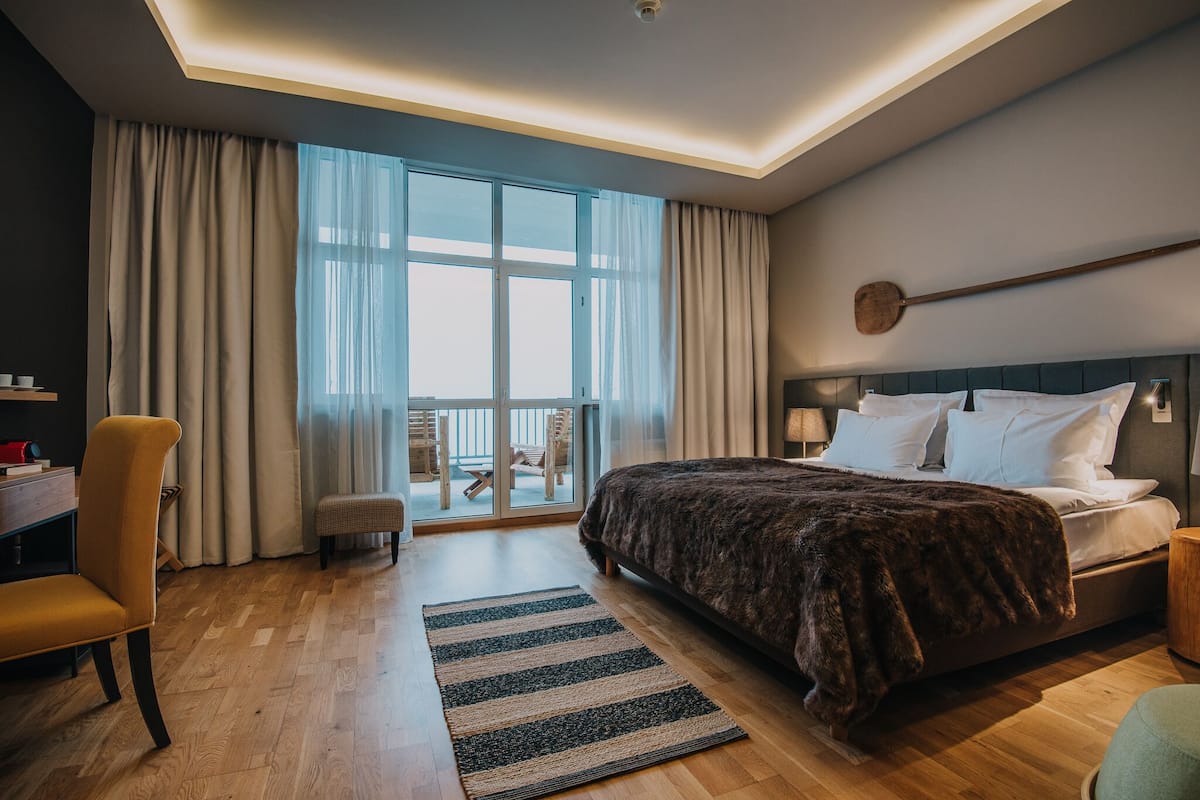 A modern hotel room with a large bed, a desk, a chair, and a balcony with a view.