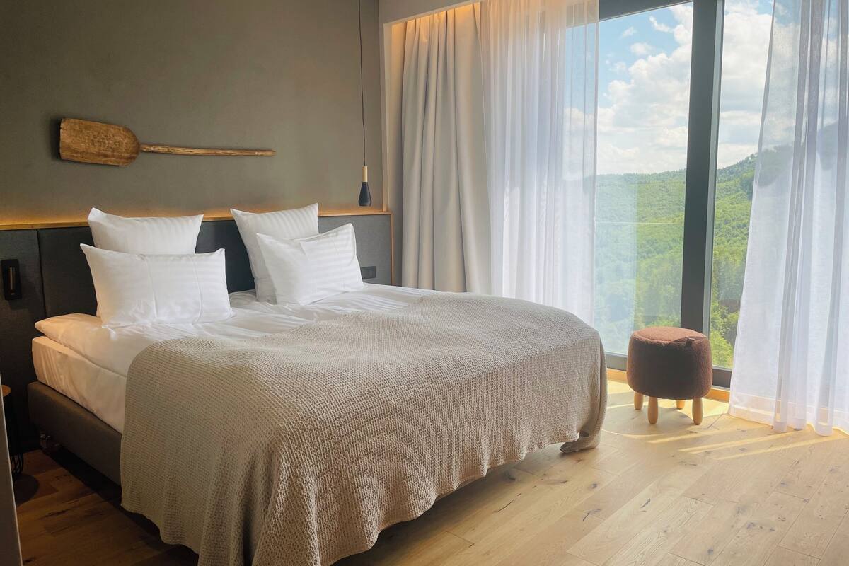 A modern bedroom with a large bed, a wooden headboard, and a view of a forest through a large window.