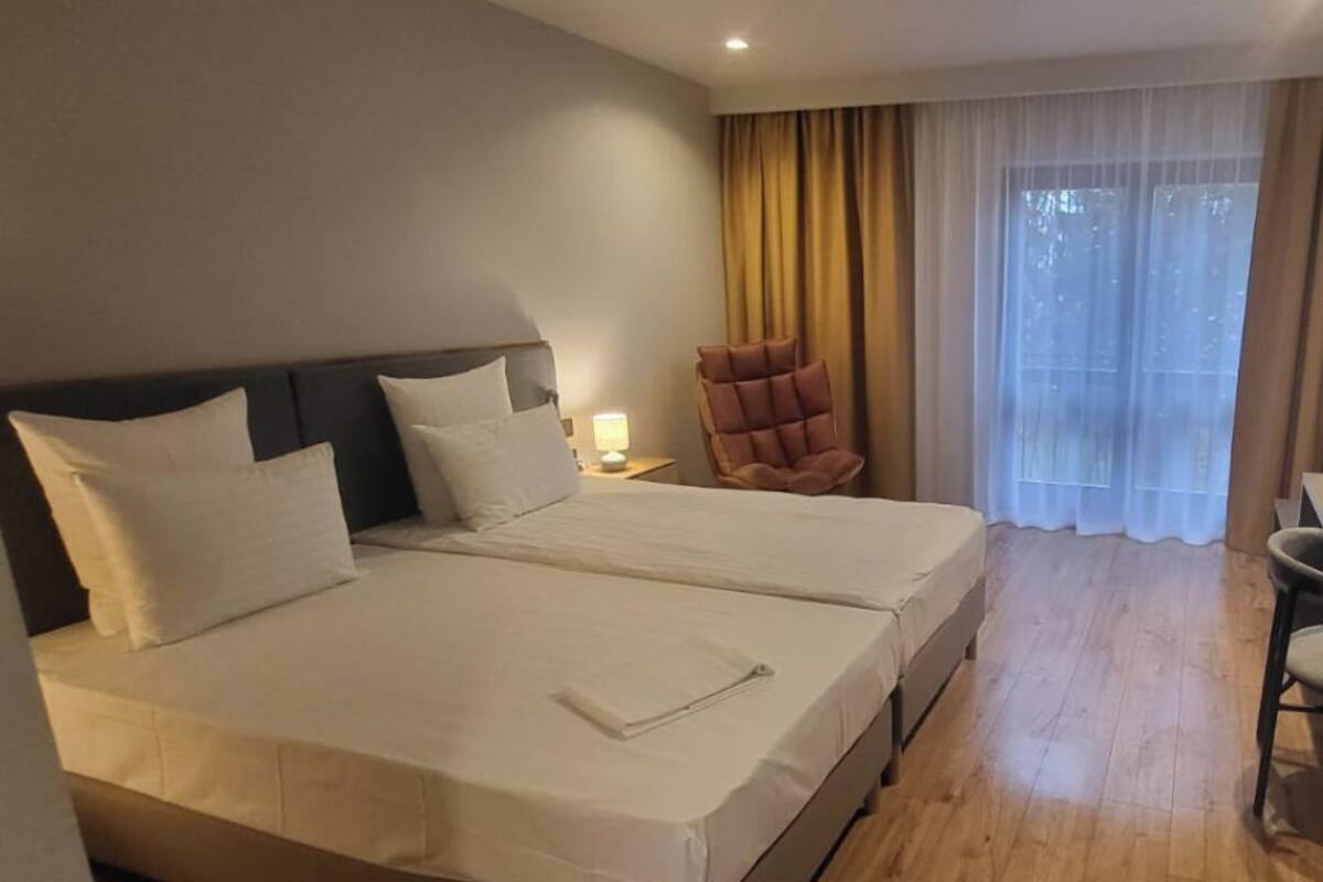 A double bed with white linens, a nightstand with a lamp, a cushioned chair, and a large window with curtains.