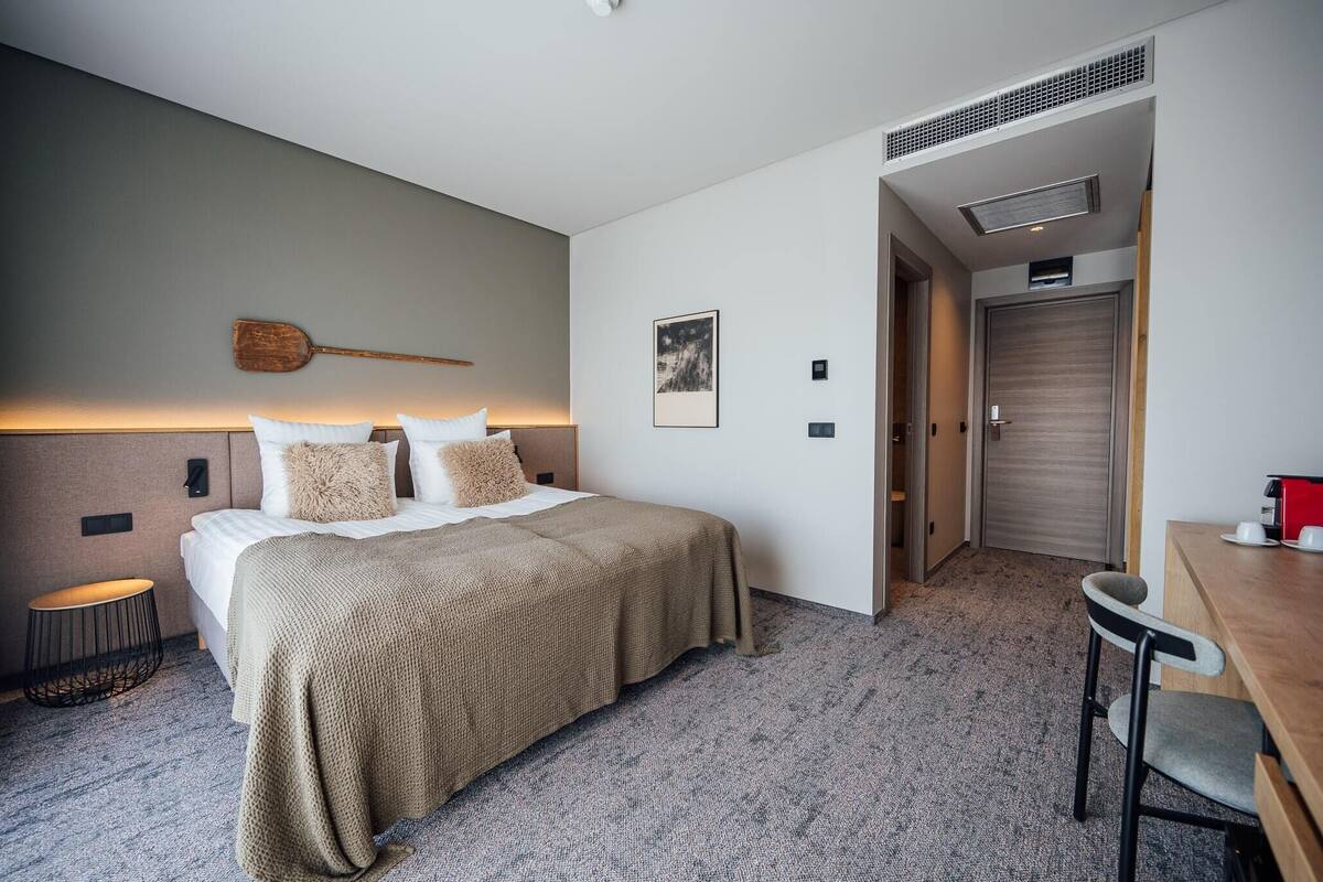A hotel room with a bed, a small round table, a chair, a wooden door, and a wall-mounted light fixture.