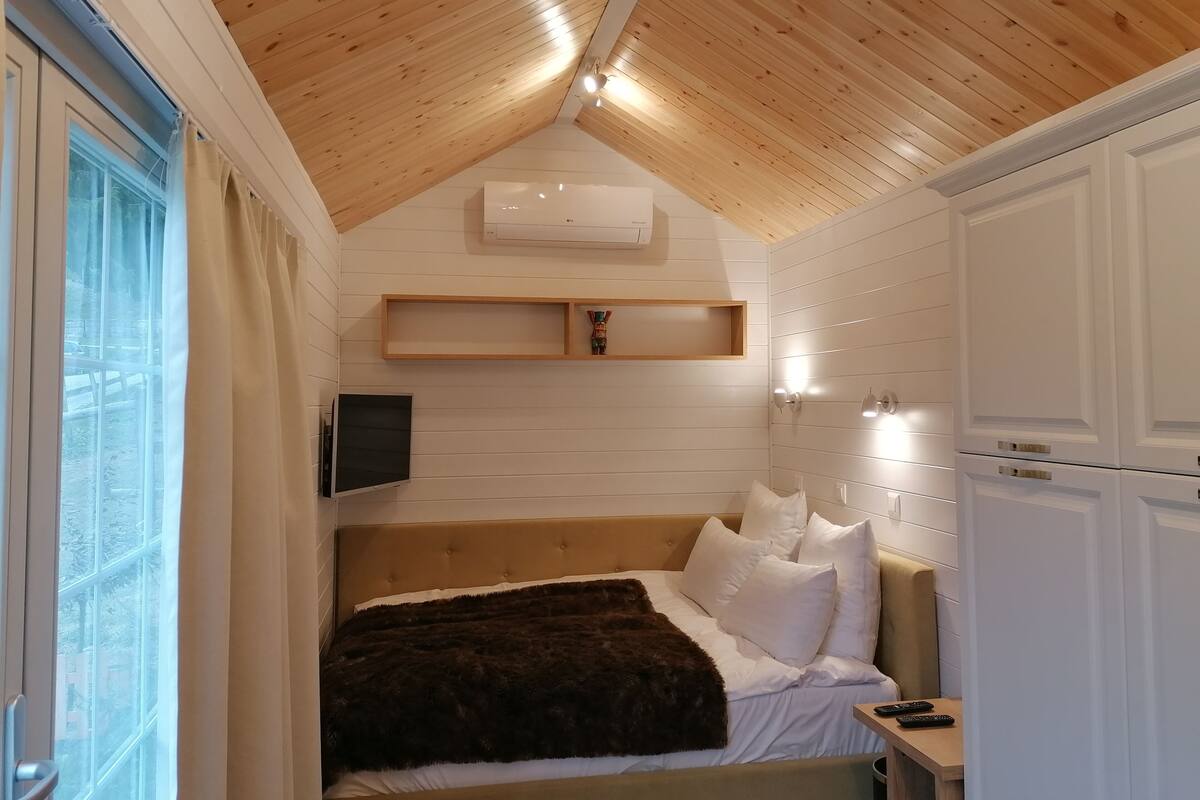 A bedroom with a wooden ceiling, a bed with white bedding, a mounted TV, and a small shelf.