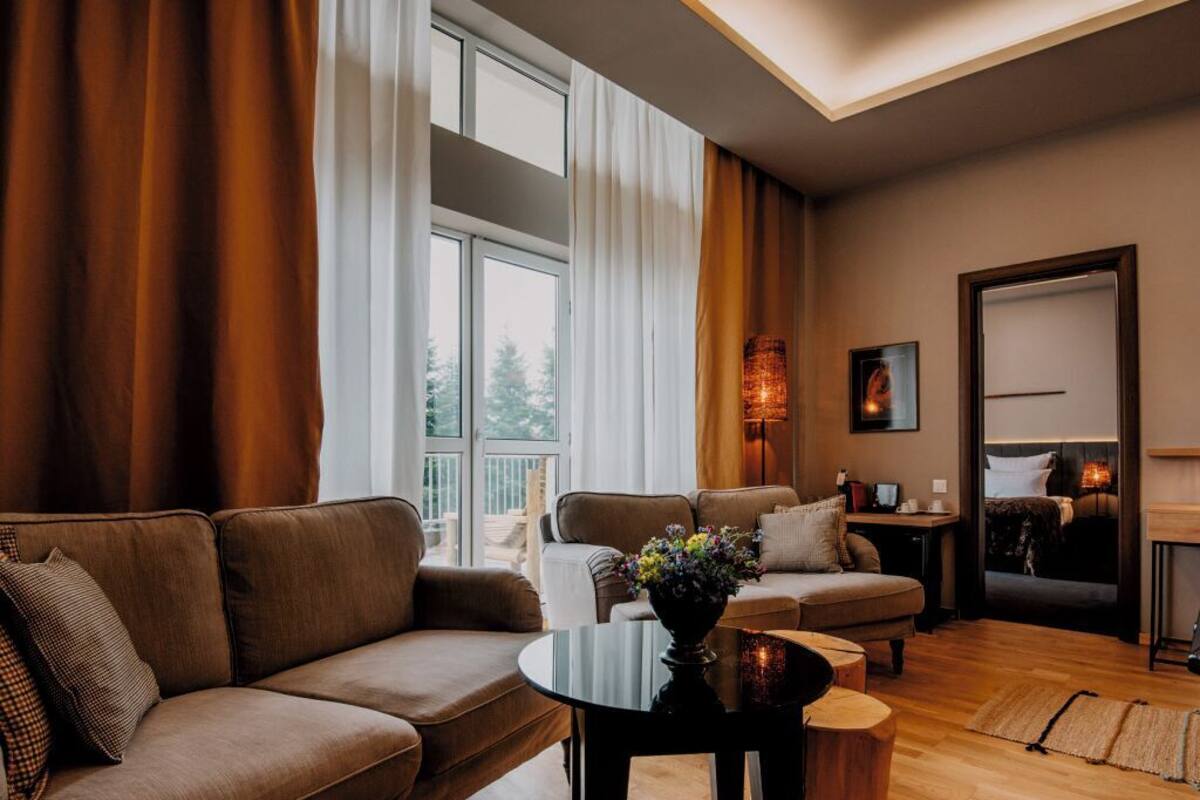 A modern living room with a brown sofa, a round coffee table, and a wooden side table. There is a window with curtains, a lamp, and a framed picture on the wall.
