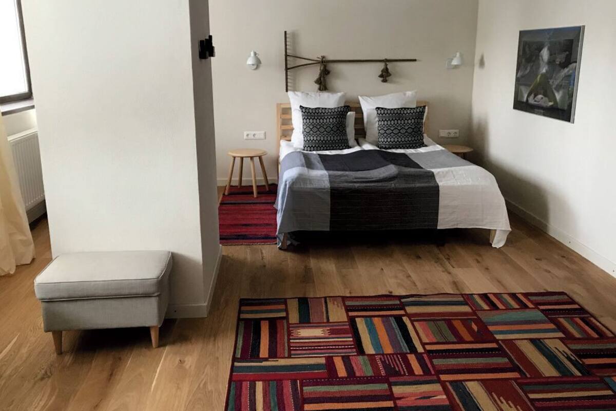 A bedroom with a bed, a nightstand, a small stool, a colorful rug, and a painting on the wall.
