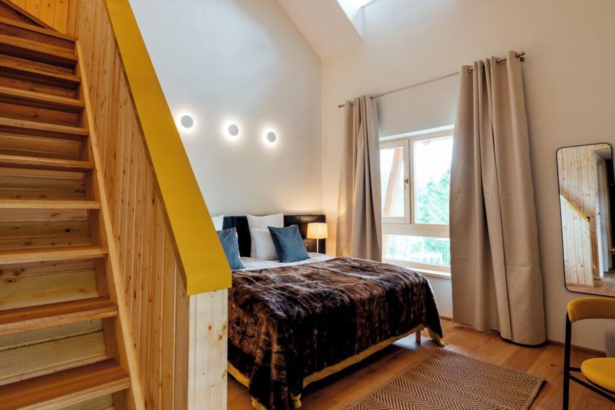 A bedroom with a wooden staircase, a bed with a patterned bedspread, a window with curtains, and a mirror on the wall.