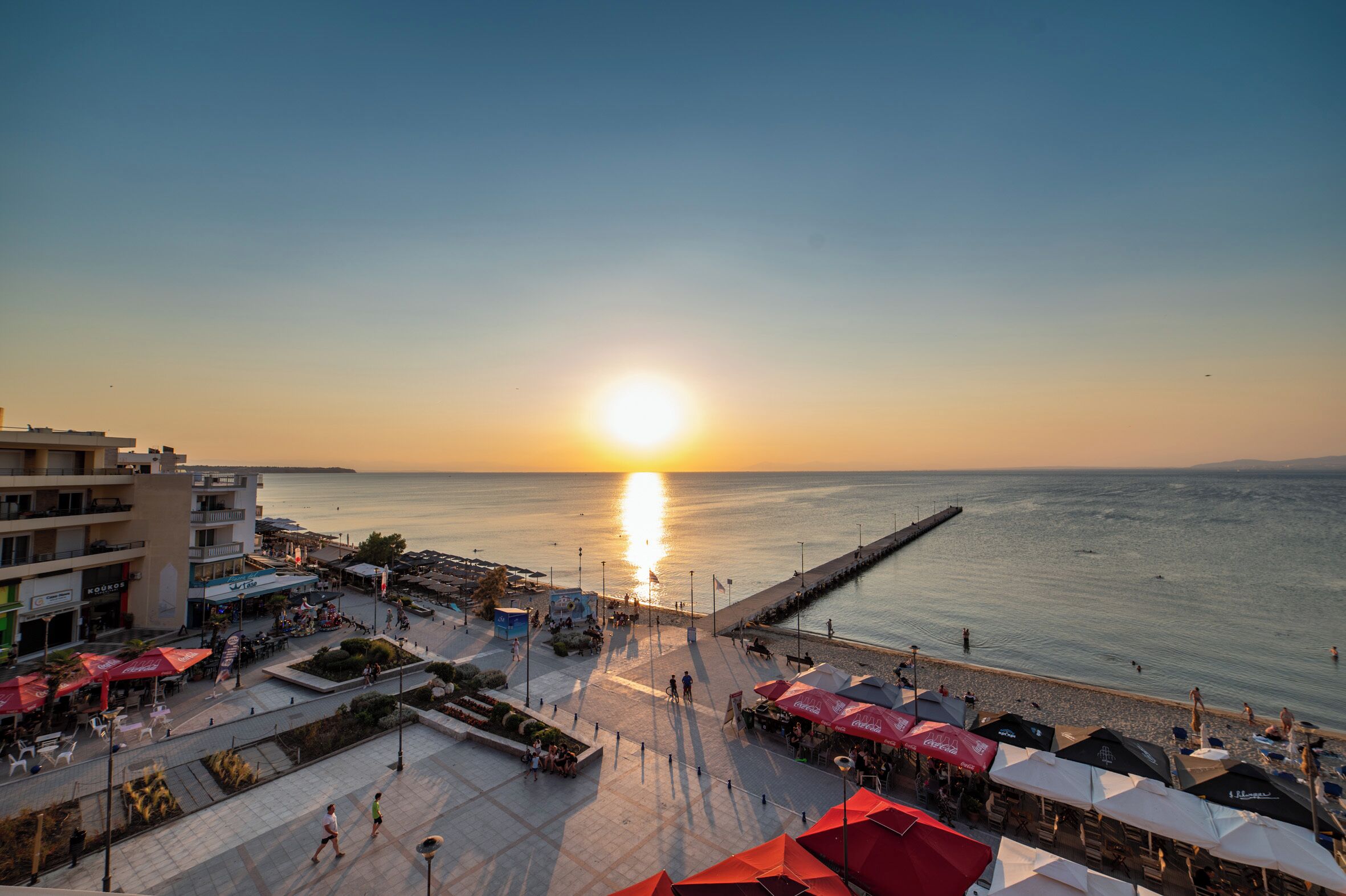 Un paseo marítimo con vistas al atardecer, mesas al aire libre y zona de restaurantes frente al agua.
