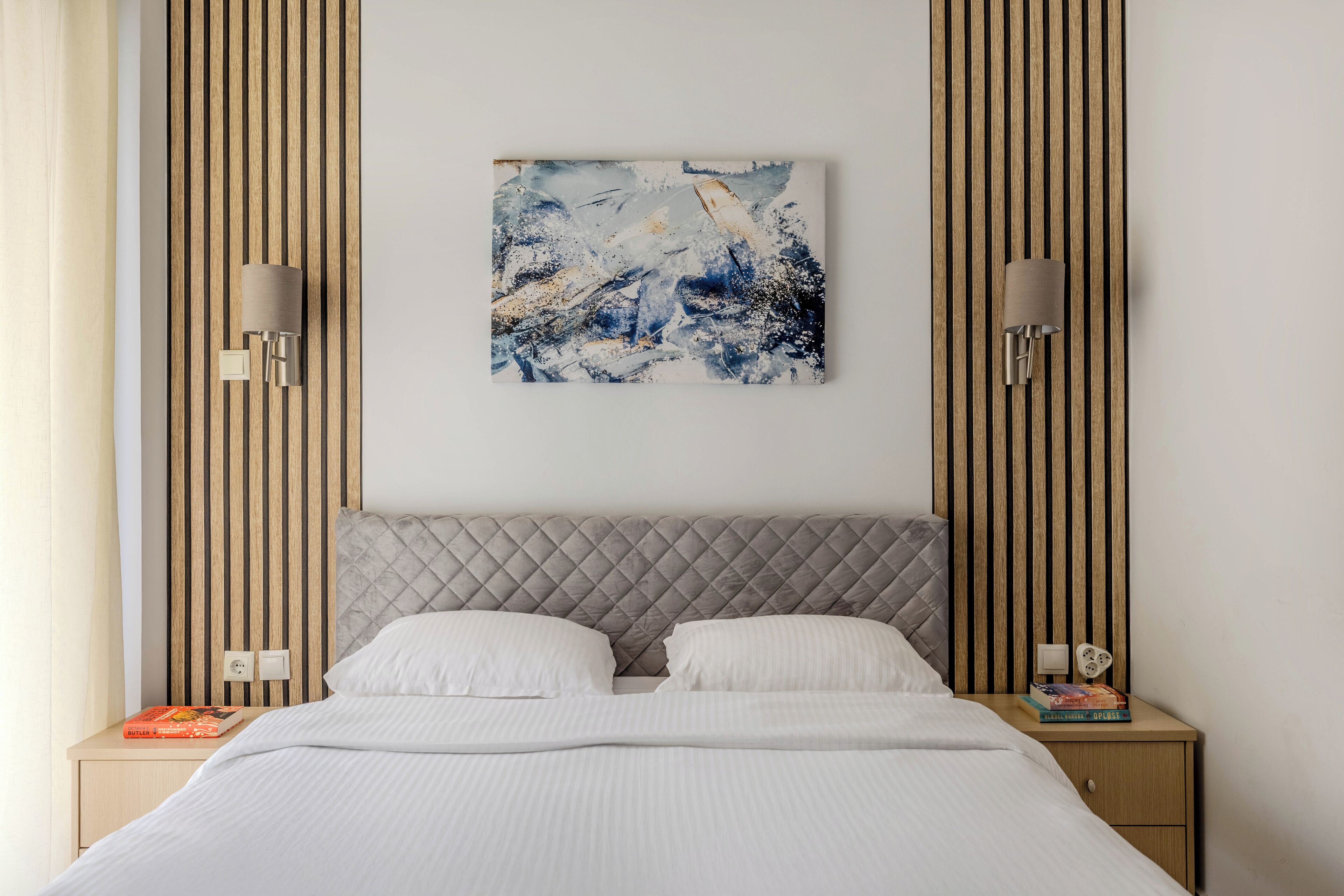 A neatly made bed with white linens, a grey quilted headboard, and a single abstract painting above it.