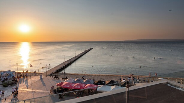 A beachside promenade with a pier extending into the water, people walking, and umbrellas set up for shade. - Vento di Mare (Thermaikos)