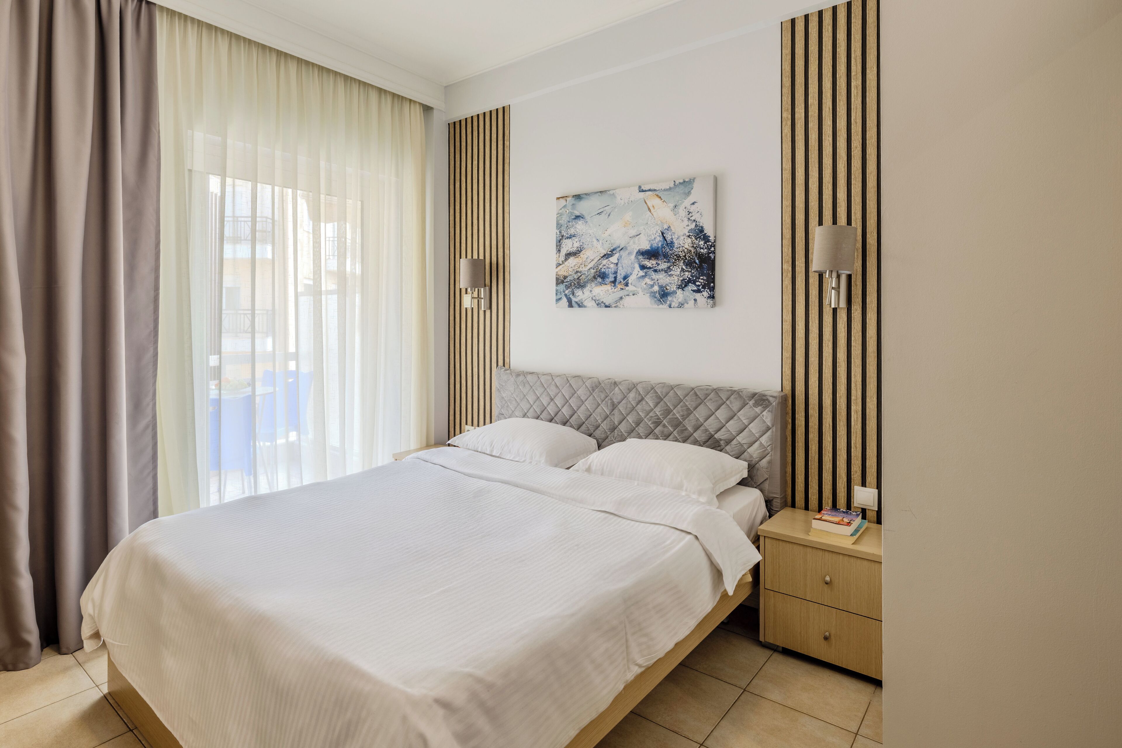 A modern hotel room with a large bed, a nightstand, a wall-mounted lamp, and a painting.