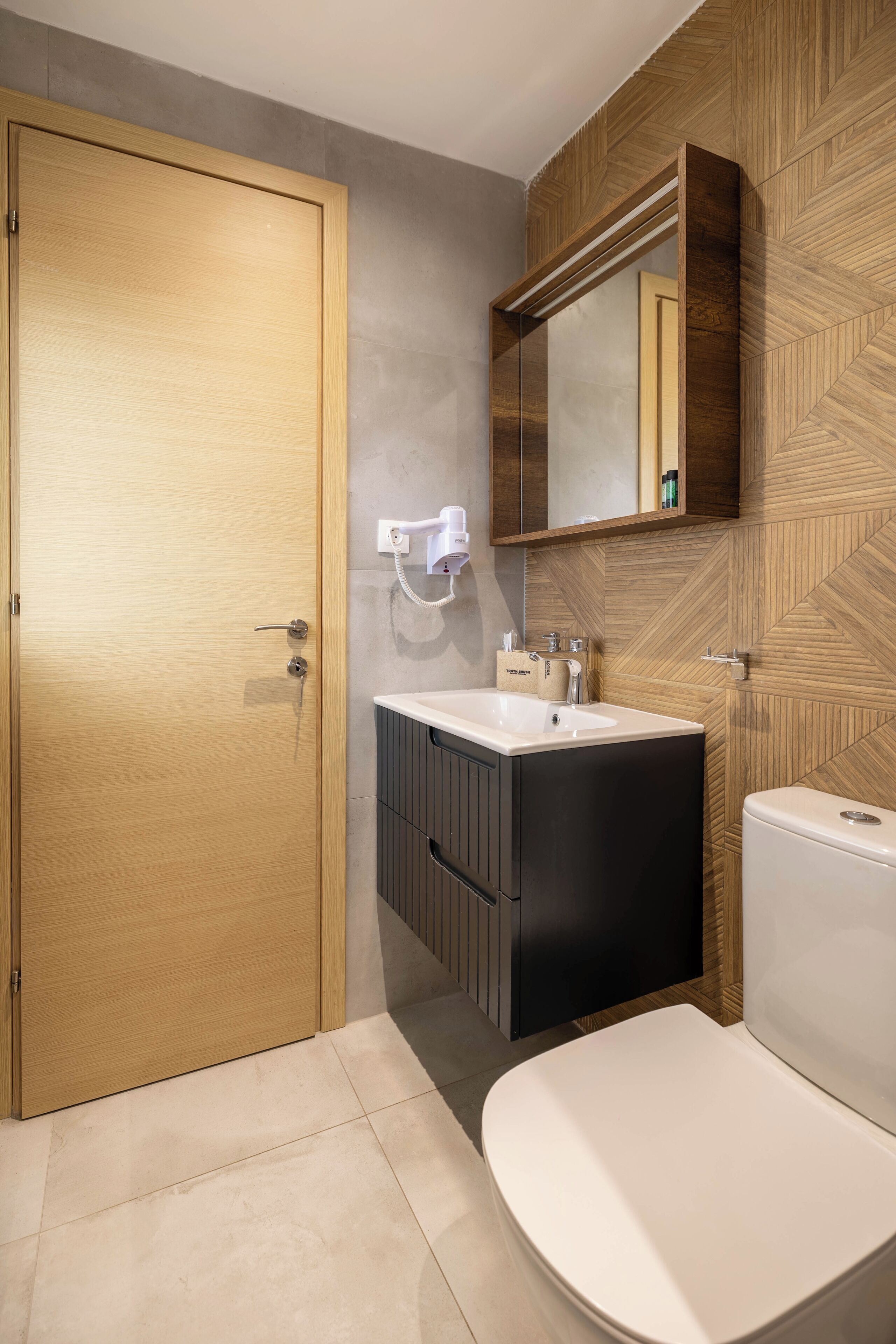 A modern bathroom with a wooden door, a white sink, a black cabinet, and a white toilet.