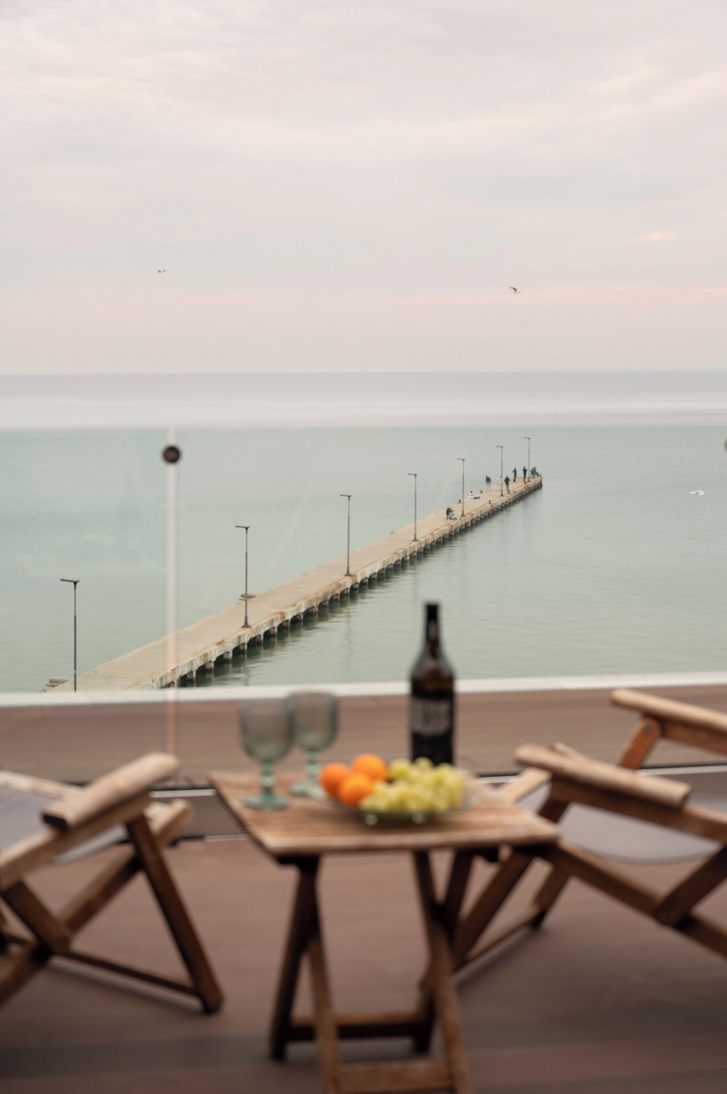 Un tavolo da pranzo con una bottiglia e della frutta, con vista su un molo che si protende in acque calme.