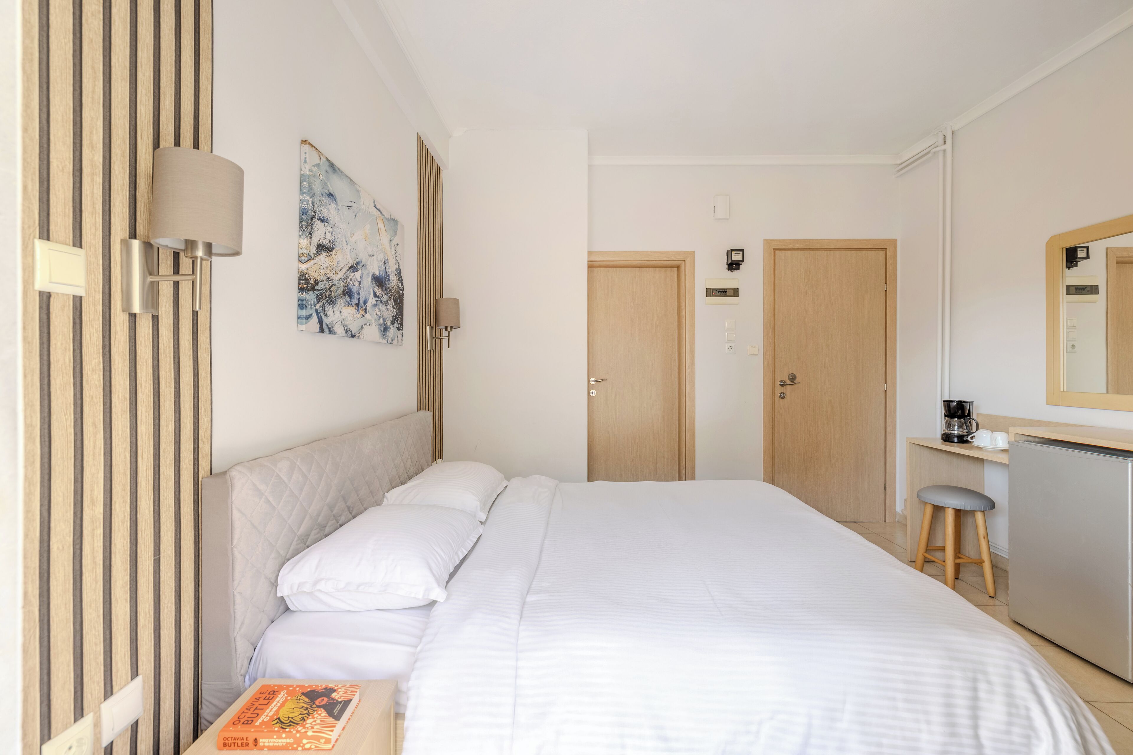 A hotel room with a bed, a desk with a chair, a coffee machine, and a mirror.