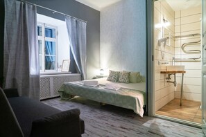 A bedroom with a bed, a window with curtains, a bedside table, and a bathroom with a shower. - Godart Rooms (Tallinn)