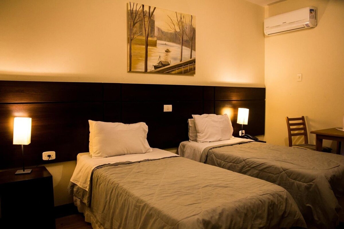 A hotel room with two beds, a nightstand, a desk, a chair, an air conditioner, and a painting on the wall.
