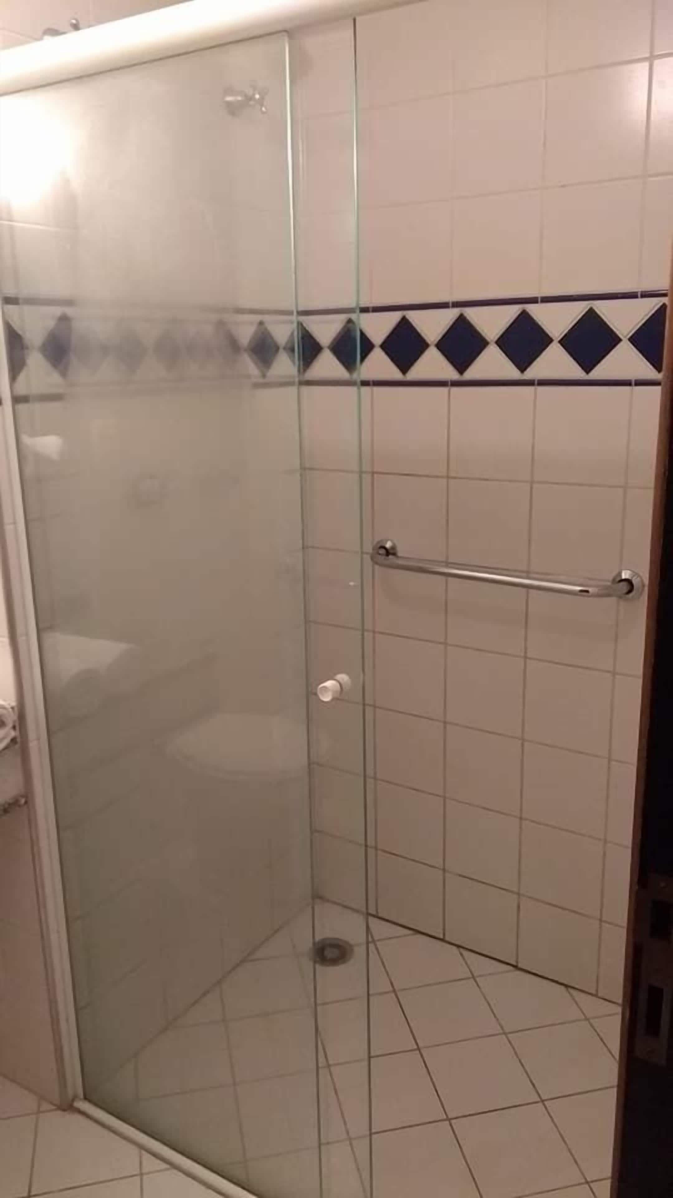 Divisória de chuveiro envidraçada com barra de apoio metálica, paredes revestidas com azulejos e uma pequena janela.