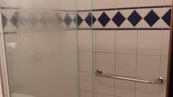 Divisória de chuveiro envidraçada com barra de apoio metálica, paredes revestidas com azulejos e uma pequena janela.