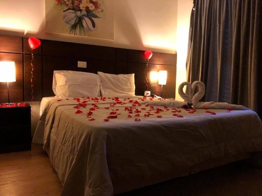 A hotel room with a large bed, two bedside lamps, a bouquet of flowers on the wall, and a heart-shaped decoration on the bed.
