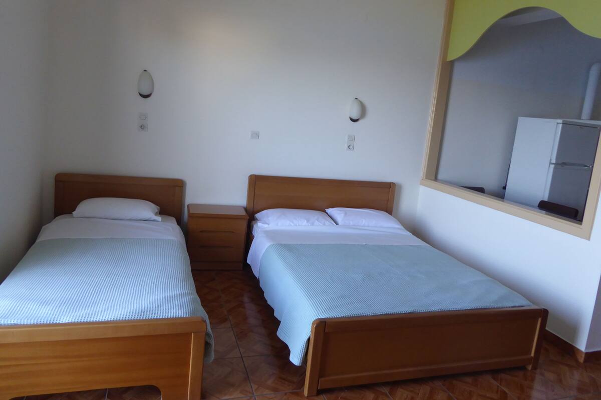 Una habitación con dos camas de madera, una mesita de noche, una lámpara de pared y una ventana con vista a un refrigerador.