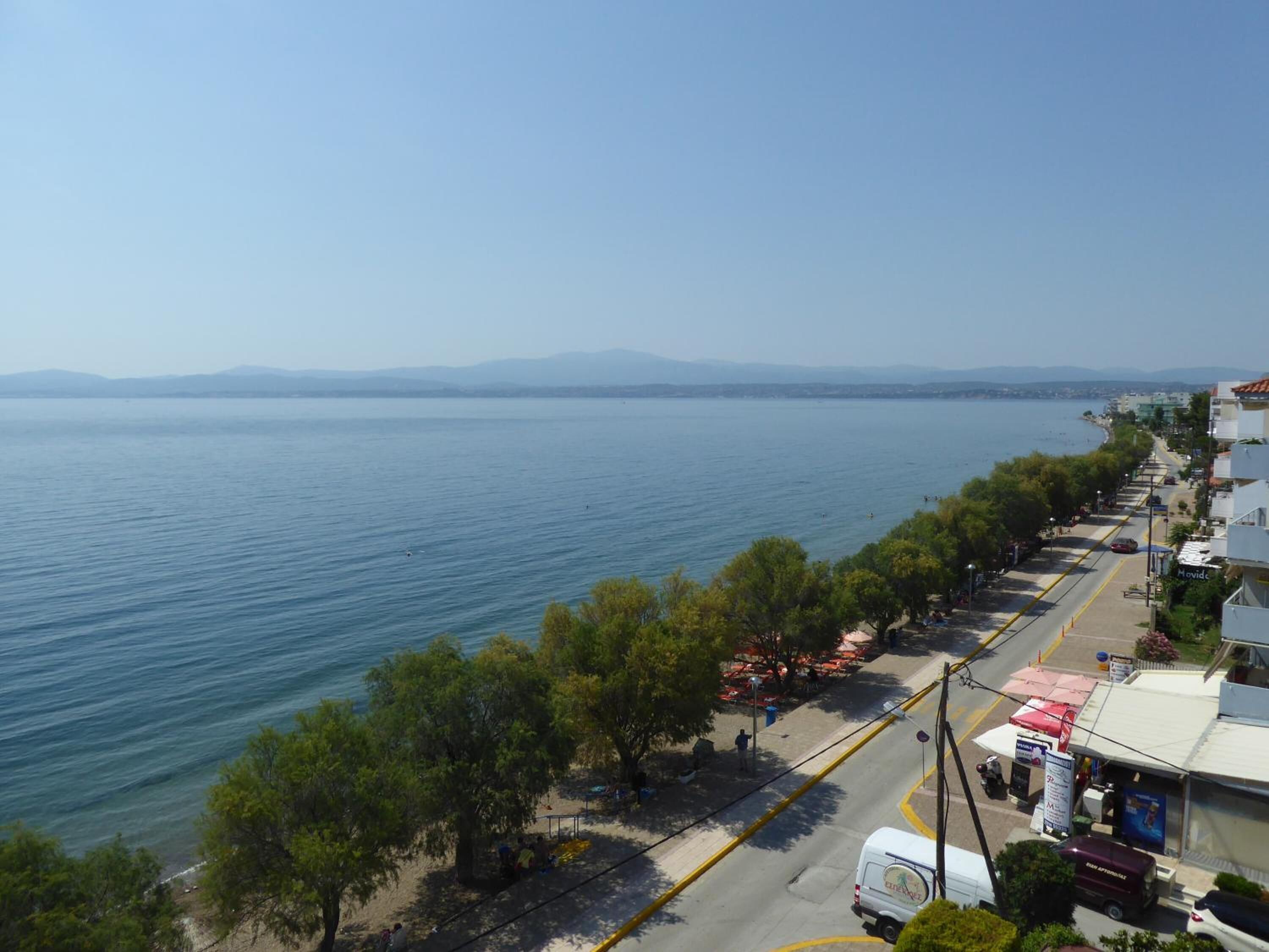 A coastal road with a view of the sea and distant mountains. There are trees lining the road, and a few people are visible walking.