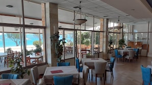 A spacious restaurant with large windows offering a view of the sea, multiple tables and chairs, and hanging pendant lights. - Hotel Poseidon (Chalcis)