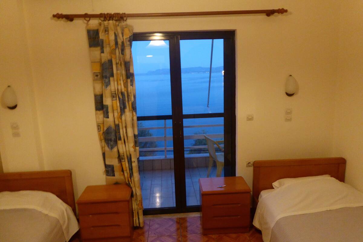 Habitación de hotel con dos camas, una cortina, vista al mar a través de una puerta corrediza y una pequeña mesa.