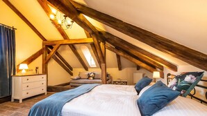 Un dormitorio con techo inclinado, vigas de madera, cama con ropa de cama azul, mesita de noche con lámpara y ventana con cortinas.