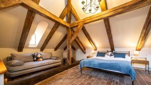 Un dormitorio con techo inclinado, vigas de madera a la vista, suelo de ladrillo, una cama con colcha azul, una mesita de noche con lámpara y un sofá con cojines.