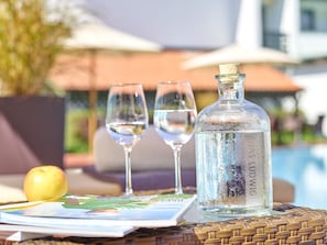 A bottle of water, two glasses, a magazine, and a fruit on a wicker table by a pool.
