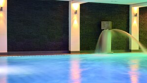 A swimming pool with a water feature, surrounded by dark brick walls and illuminated by wall sconces. - DAS LUDWIG Sports Resort (Bad Griesbach im Rottal)