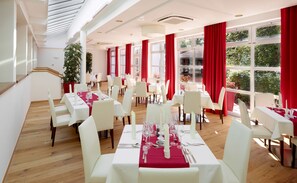 A dining area with white tables and chairs, red curtains, and large windows with a view of greenery.