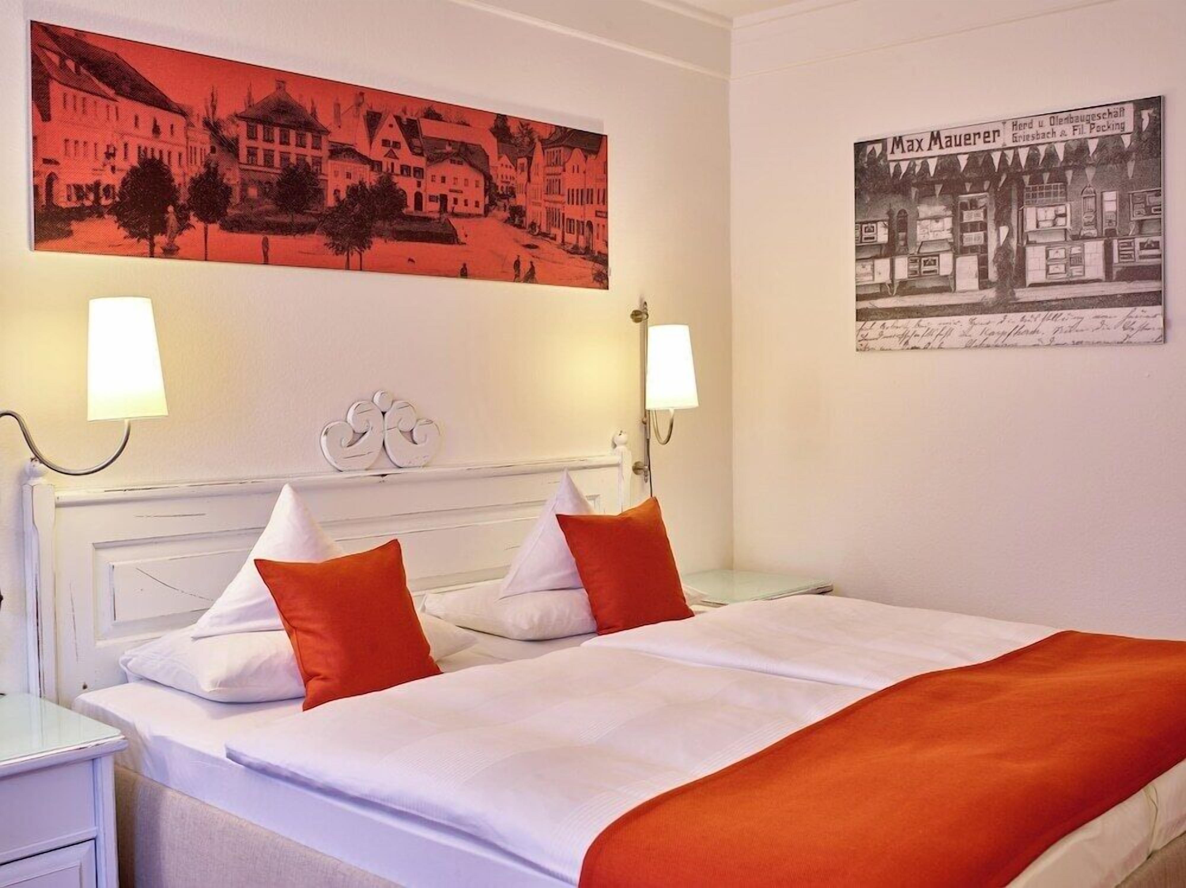 A hotel room with a bed, two bedside lamps, a headboard with a crest, and two paintings on the wall.