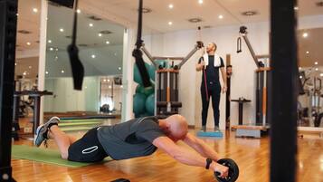 Ett gym med träningsutrustning, en person som gör en planka på en matta och en annan person som står och använder ett träningsband.