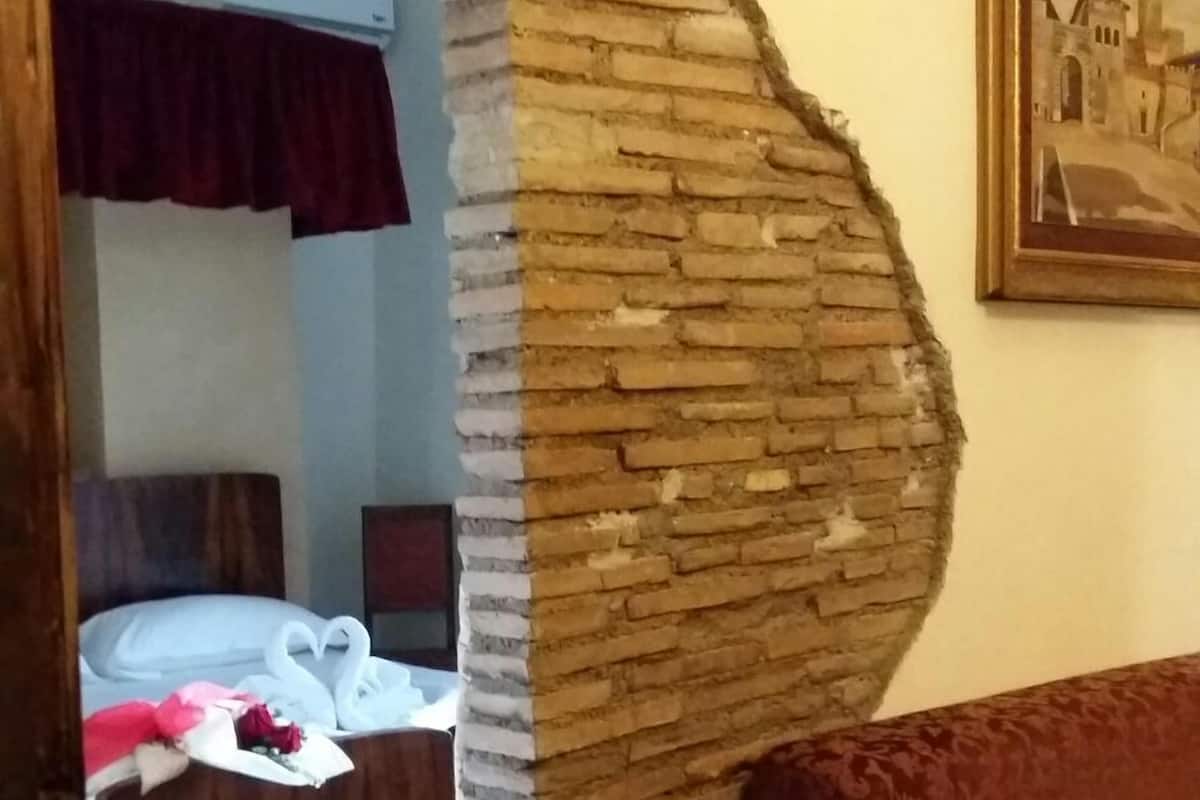 Ein Zimmer mit einer teilweise eingestürzten Backsteinmauer, einem Bett mit einer schwanen-förmigen Dekoration, einem roten Vorhang und einem gerahmten Bild an der Wand.