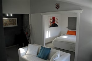 Un dormitorio moderno con cama blanca, almohada naranja decorativa y sofá blanco con cojín azul. Hay un cuadro en la pared y una mesita con una silla.