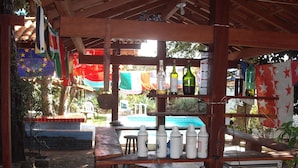 A rustic outdoor bar area with wooden beams, a pool in the background, and various bottles and glasses on shelves. - Pousada Chalé Suiço (Parnaíba)