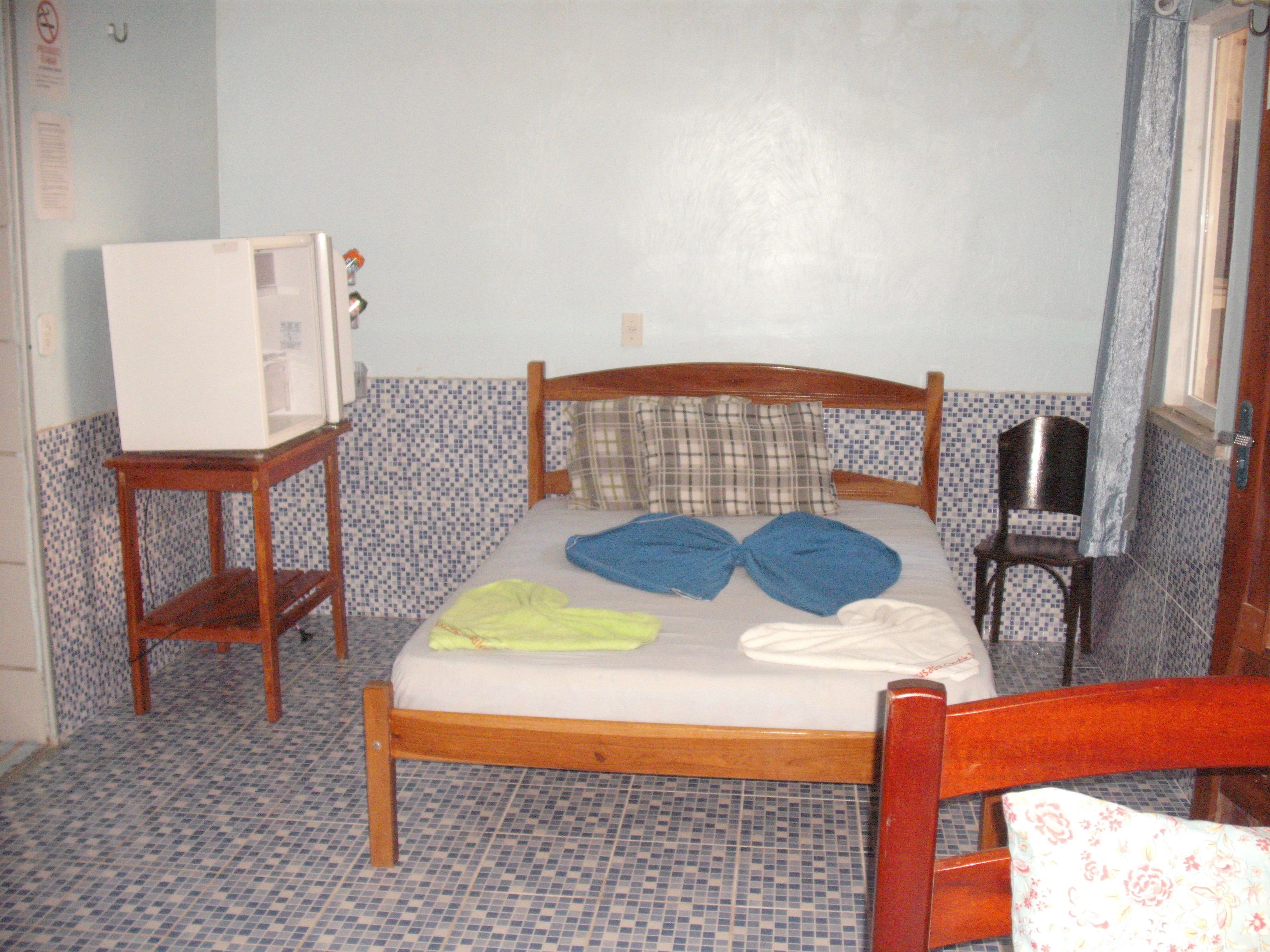 A small, simple room with a bed, a wooden chair, a small table, and a television on a stand.