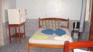 A small, simple room with a bed, a wooden chair, a small table, and a television on a stand.